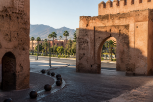 Porte ocre de Marrakech ouverte sur une avenue arborée et les reliefs de l’Atlas